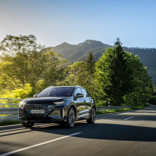Audi Q4 SUV e-tron in Tamboragrey, outdoor driving shot, exterior, three-quarter front view on a country road surrounded by forest and mountain scenery.
